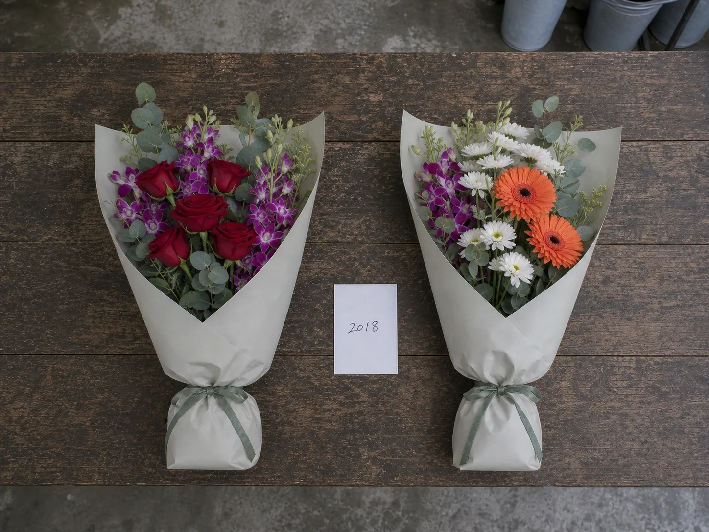 Two wrapped bouquets of the same size lying side by side on the shop bench — red roses with purple orchid, and orange gerbera with white chrysanthemum — with a handwritten card reading 2018 between them