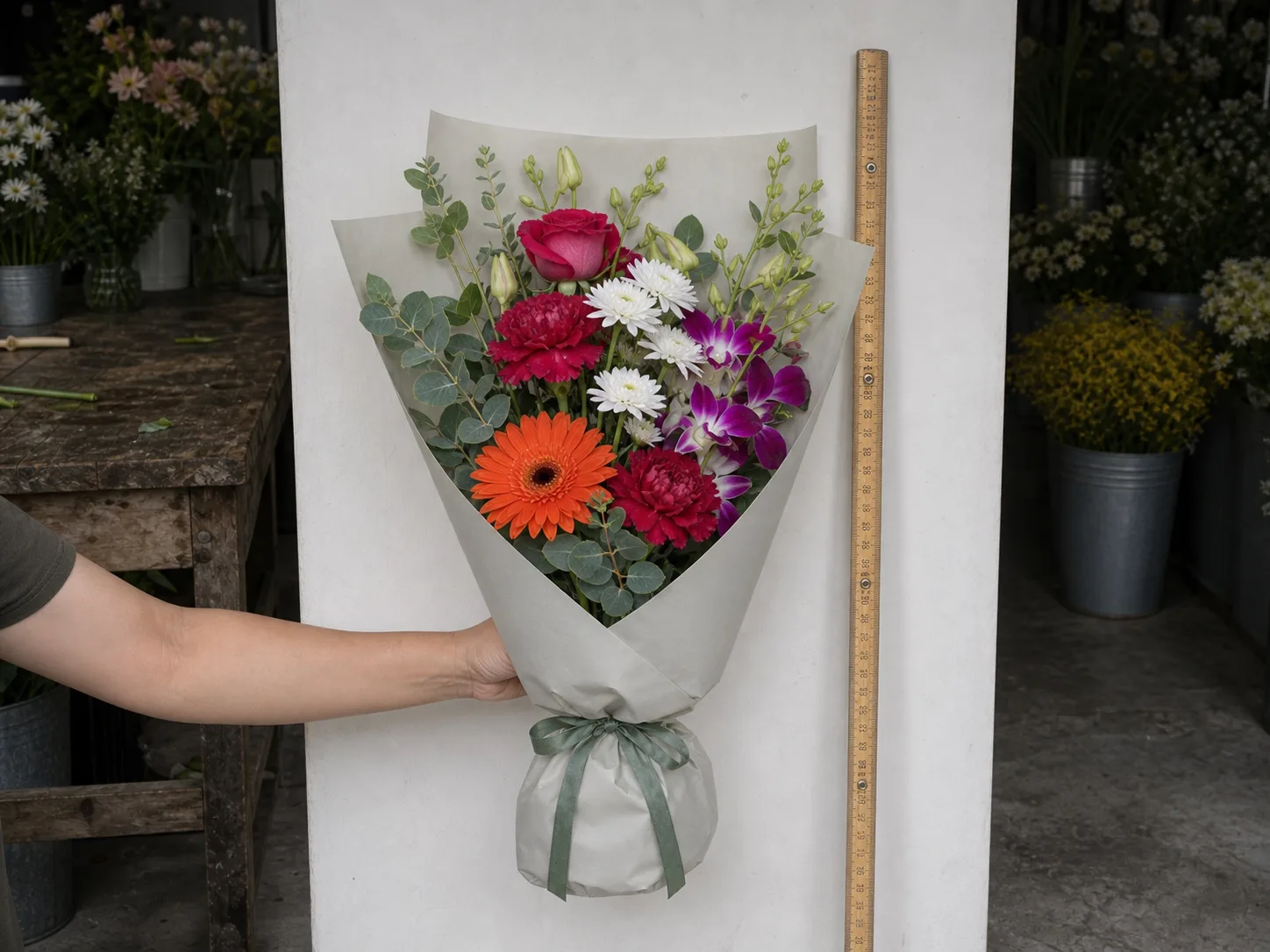 A mixed birthday bouquet in orange, pink and purple, about 35 cm across, held upright against a plain wall beside a ruler