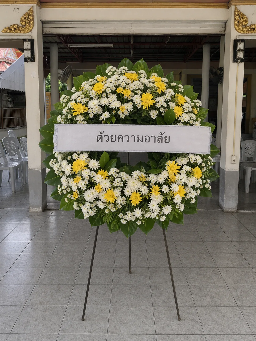 A standing funeral wreath of white and yellow chrysanthemum on a tripod frame, its ribbon reading ด้วยความอาลัย, set up at a temple sala before the evening